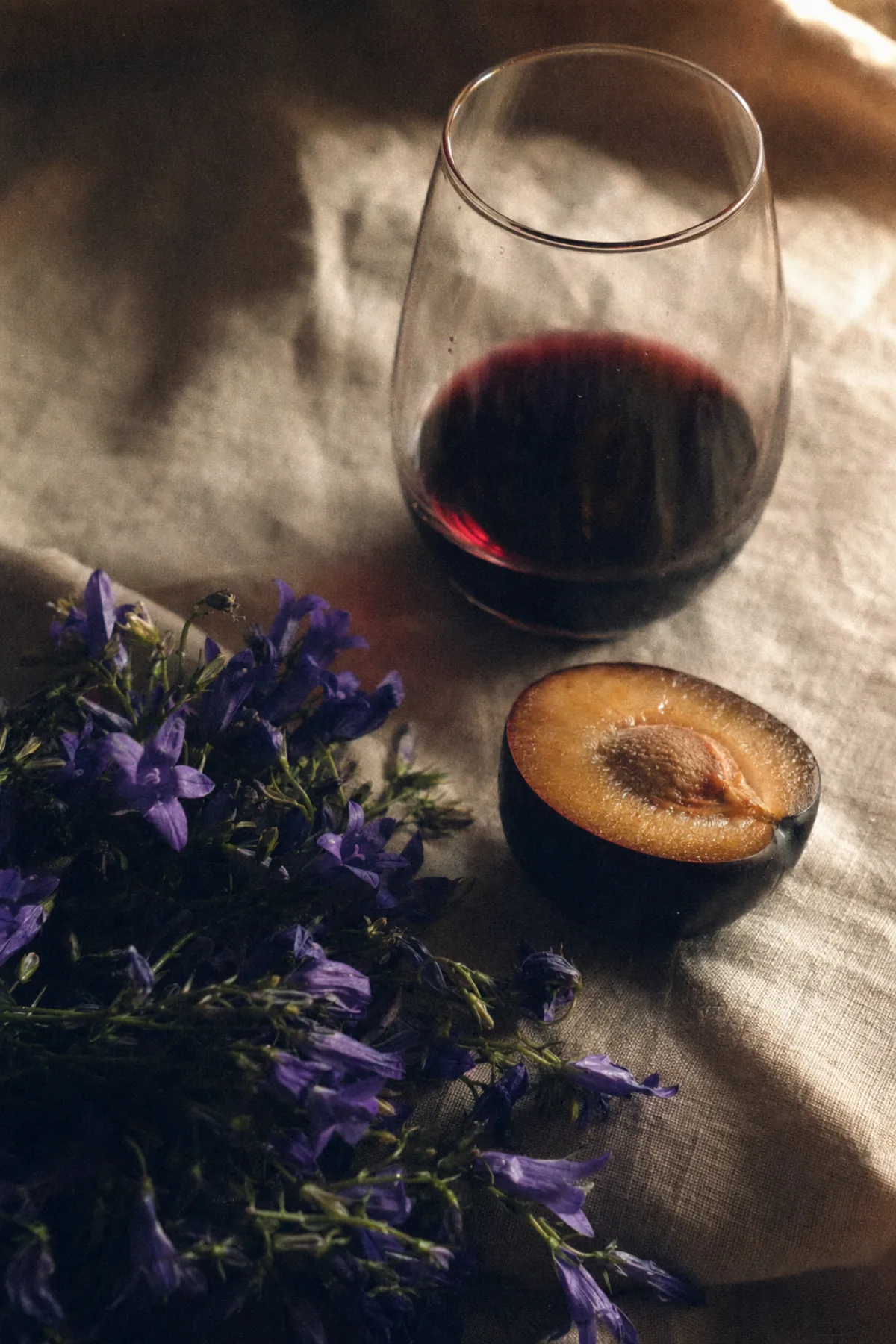 Violet flowers, a ripe black plum cut in half, and a stemless pour of marselan on linen