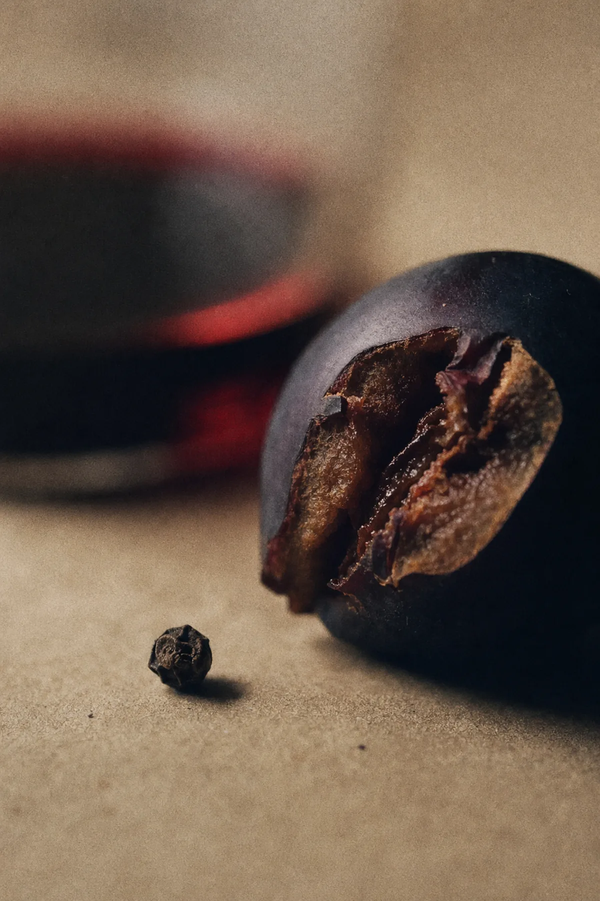 A black peppercorn on craft paper beside a smoked plum, a pour of shiraz out of focus